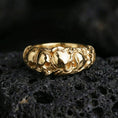 Load image into Gallery viewer, Gold plated skull band ring on textured volcanic rock, premium close-up with warm reflective shine across the skull reliefs and raised sculpted surfaces.