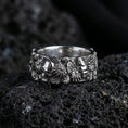 Load image into Gallery viewer, Polished silver skull and floral band ring on textured volcanic rock, crisp macro product photo emphasizing clean edges, sharp petal detailing, and bright reflective highlights across the sculpted band.