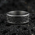 Load image into Gallery viewer, Oxidized geometric knotwork band ring on textured volcanic rock, centered close-up highlighting the repeating interlace pattern, carved depth, and deep antique shadowed contrast.