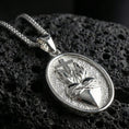 Load image into Gallery viewer, Polished silver Christian medal with Sacred Heart motif, radiant flame relief, and smooth oval frame placed on textured volcanic rock in a premium studio product shot