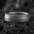 Load image into Gallery viewer, Oxidized Damascus-style band ring on volcanic stone, macro close-up highlighting darkened layered wave lines and brushed edges.