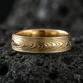 Load image into Gallery viewer, Gold plated Damascus-style band ring on volcanic stone, premium close-up showing warm gold sheen over the layered grain pattern and smooth borders.