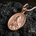 Load image into Gallery viewer, Rose gold plated oval Saint Jude necklace with haloed portrait, devotional relief, and sacred medallion detail resting on volcanic rock in an elegant close-up product presentation