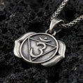 Load image into Gallery viewer, Oxidized third eye chakra pendant with Om symbol, triangle motif, and two-petal lotus design displayed on dark volcanic rock in a close-up luxury jewelry composition
