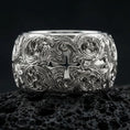 Load image into Gallery viewer, Polished silver ornate cross band ring on volcanic stone, crisp product photo emphasizing the bright reflective finish and detailed baroque engravings.