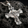 Load image into Gallery viewer, Polished silver Archangel Michael cross pendant with raised warrior relief, shield detail, and dramatic winged silhouette placed on textured volcanic rock in a premium studio product shot