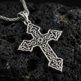 Load image into Gallery viewer, Oxidized Celtic cross pendant with intricate knotwork details and open ornamental silhouette displayed on dark volcanic rock in a close-up luxury jewelry composition