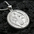 Load image into Gallery viewer, Polished silver Saint Benedict medal with detailed saint relief, engraved Benedictine symbols, and circular frame placed on textured volcanic rock in a premium studio product shot