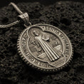 Load image into Gallery viewer, Bronze Saint Benedict medallion pendant with robed saint figure, Latin inscriptions, and round devotional design displayed on dark volcanic rock in a close-up luxury jewelry composition