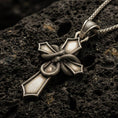 Load image into Gallery viewer, Bronze cross pendant with wrapped snake detail and bold gothic silhouette displayed on dark volcanic rock in a close-up luxury jewelry composition