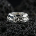 Load image into Gallery viewer, Polished silver mountain scene band ring on black volcanic stone, crisp raised peak engraving with natural terrain detail, centered close-up studio lighting.
