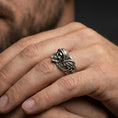 Load image into Gallery viewer, Oxidized sterling silver biker skull ring worn on finger, helmeted skull relief with wing accents, natural lifestyle hand shot with shallow depth of field.