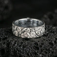 Load image into Gallery viewer, Polished silver floral engraved band ring on black volcanic stone, crisp daisy relief and swirling botanical pattern, centered close-up studio lighting.