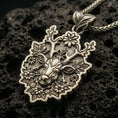 Load image into Gallery viewer, Bronze deer pendant with floral antlers, detailed stag face, and nature-inspired silhouette displayed on dark volcanic rock in a close-up luxury jewelry composition