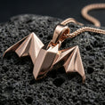 Load image into Gallery viewer, Rose gold plated bat pendant with angular origami wings, minimalist gothic form, and elegant sculptural silhouette resting on volcanic rock in an elegant close-up product presentation