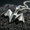 Load image into Gallery viewer, Polished silver bat necklace with angular origami-inspired form, faceted wing structure, and modern gothic animal design placed on textured volcanic rock in a premium studio product shot