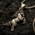Load image into Gallery viewer, Bronze horse pendant with rearing equine silhouette, flowing mane, and detailed muscular form displayed on dark volcanic rock in a close-up luxury jewelry composition