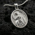 Load image into Gallery viewer, Polished silver religious medal pendant with a seated angel relief and oval border placed on textured volcanic rock in a premium studio product shot