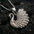 Load image into Gallery viewer, Polished silver swan necklace with layered feather detailing, sculpted wing fan design, and refined bird motif placed on textured volcanic rock in a premium studio product shot