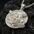 Load image into Gallery viewer, Polished silver Saint George medallion with detailed horse and dragon relief, Corinthians banner, and circular frame placed on textured volcanic rock in a premium studio product shot