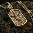 Load image into Gallery viewer, Bronze Jesus pendant with ascending Christ figure, angel motifs, and framed devotional design displayed on dark volcanic rock in a close-up luxury jewelry composition
