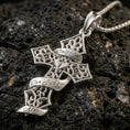 Load image into Gallery viewer, Polished silver Christian cross pendant with openwork gothic pattern, engraved ribbon text, and decorative cross silhouette placed on textured volcanic rock in a premium studio product shot