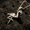 Load image into Gallery viewer, Bronze origami flamingo pendant necklace with an oxidized finish resting on textured volcanic rock, captured in a close-up premium studio composition.