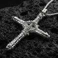 Load image into Gallery viewer, Polished silver gothic cross necklace with biomechanical relief, layered structural textures, and elongated cross silhouette placed on textured volcanic rock in a premium studio product shot