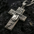 Load image into Gallery viewer, Oxidized Jesus cross pendant with sculpted crucifix figure, crown of thorns, and textured relief displayed on dark volcanic rock in a close-up luxury jewelry composition