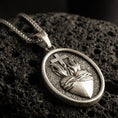 Load image into Gallery viewer, Bronze Sacred Heart pendant with cross, flame detail, and oval devotional design displayed on dark volcanic rock in a close-up luxury jewelry composition