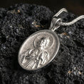 Load image into Gallery viewer, Polished silver Saint Jude medal with oval frame, sacred portrait relief, and halo detail placed on textured volcanic rock in a premium studio product shot