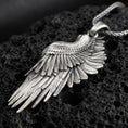 Load image into Gallery viewer, Oxidized angel wing pendant with layered feather detailing and elongated wing silhouette displayed on dark volcanic rock in a close-up luxury jewelry composition