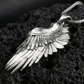 Load image into Gallery viewer, Polished silver wing necklace with sculpted feather texture, curved angel wing form, and refined dimensional relief placed on textured volcanic rock in a premium studio product shot