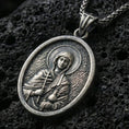 Load image into Gallery viewer, Oxidized Saint Anastasia pendant with haloed saint figure, cross detail, and oval devotional portrait displayed on dark volcanic rock in a close-up luxury jewelry composition