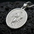 Load image into Gallery viewer, Polished silver Saint Paul medallion with engraved saint relief, halo motif, and detailed oval frame placed on textured volcanic rock in a premium studio product shot