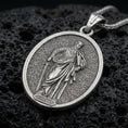Load image into Gallery viewer, Oxidized Saint Paul pendant with haloed saint figure, sword detail, and oval devotional portrait displayed on dark volcanic rock in a close-up luxury jewelry composition