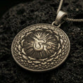 Load image into Gallery viewer, Bronze Om pendant with circular mandala design, radiant center, and layered sacred pattern displayed on dark volcanic rock in a close-up luxury jewelry composition