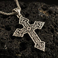 Load image into Gallery viewer, Bronze Celtic cross pendant with intricate knotwork details and open ornamental silhouette displayed on dark volcanic rock in a close-up luxury jewelry composition