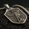 Load image into Gallery viewer, Bronze Saint Michael shield pendant necklace with an oxidized finish resting on textured volcanic rock, captured in a close-up premium studio composition.