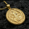 Load image into Gallery viewer, Gold plated Saint Benedict pendant with haloed saint figure, sacred lettering, and classic round medallion form showcased on black volcanic rock in a refined macro jewelry image