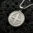 Load image into Gallery viewer, Polished silver Saint Benedict pendant with detailed cross emblem, Latin initials, and circular religious medal form placed on textured volcanic rock in a premium studio product shot
