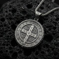 Load image into Gallery viewer, Oxidized Saint Benedict medal pendant with central cross, engraved initials, and round protective medallion design displayed on dark volcanic rock in a close-up luxury jewelry composition