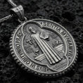 Load image into Gallery viewer, Oxidized Saint Benedict medallion pendant with robed saint figure, Latin inscriptions, and round devotional design displayed on dark volcanic rock in a close-up luxury jewelry composition