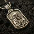 Load image into Gallery viewer, Bronze Virgin Mary and Baby Jesus pendant with halo details, framed devotional portrait, and rounded rectangular icon design displayed on dark volcanic rock in a close-up luxury jewelry composition