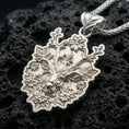 Load image into Gallery viewer, Polished silver stag necklace with blooming flower details, branched antlers, and sculpted woodland animal relief placed on textured volcanic rock in a premium studio product shot
