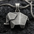 Load image into Gallery viewer, Oxidized origami elephant pendant with geometric folded form and minimalist animal silhouette displayed on dark volcanic rock in a close-up luxury jewelry composition