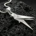 Load image into Gallery viewer, Polished silver parrot necklace with minimalist origami-inspired structure, faceted folded lines, and elongated bird shape placed on textured volcanic rock in a premium studio product shot