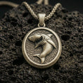 Load image into Gallery viewer, Bronze shark pendant with fierce open-mouth shark relief, oval medallion frame, and textured ocean-inspired background displayed on dark volcanic rock in a close-up luxury jewelry composition