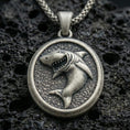 Load image into Gallery viewer, Oxidized shark pendant with fierce open-mouth shark relief, oval medallion frame, and textured ocean-inspired background displayed on dark volcanic rock in a close-up luxury jewelry composition