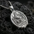 Load image into Gallery viewer, Polished silver peacock necklace with ornate plumage detailing, botanical background elements, and elegant oval frame placed on textured volcanic rock in a premium studio product shot